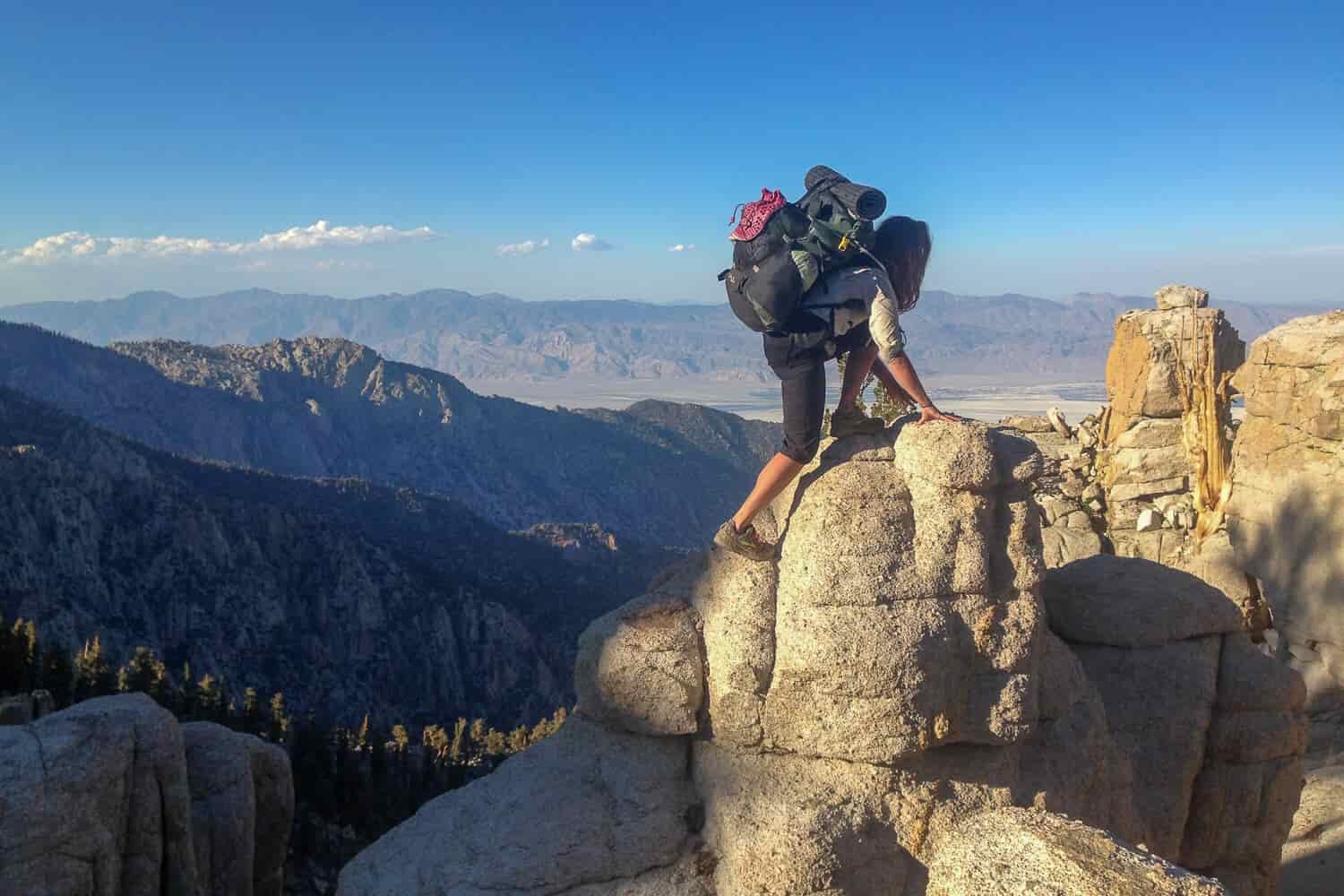 A female backpacker scrambles up weathered rocks to look down on the desert below