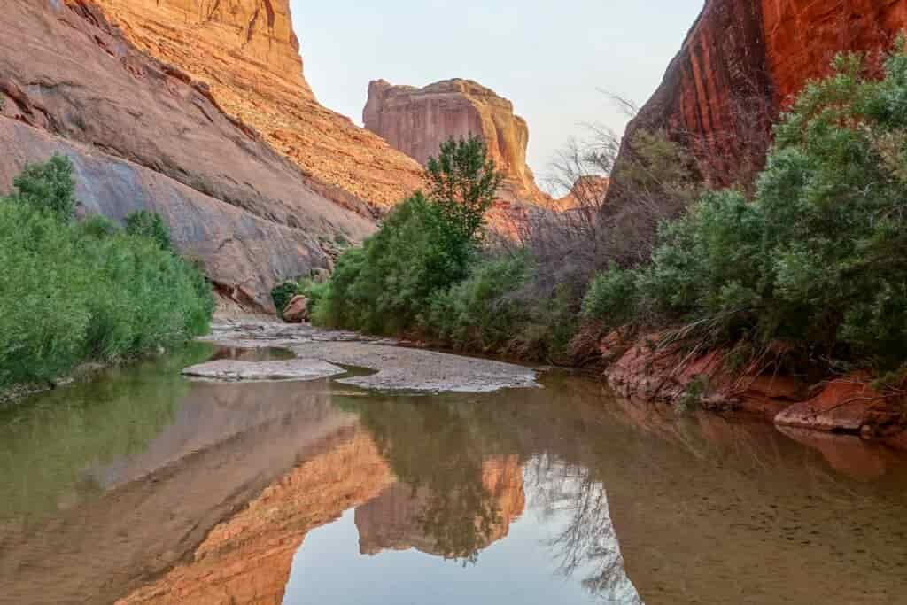 a calm body of water relfects the large sandstone rock formations behind it