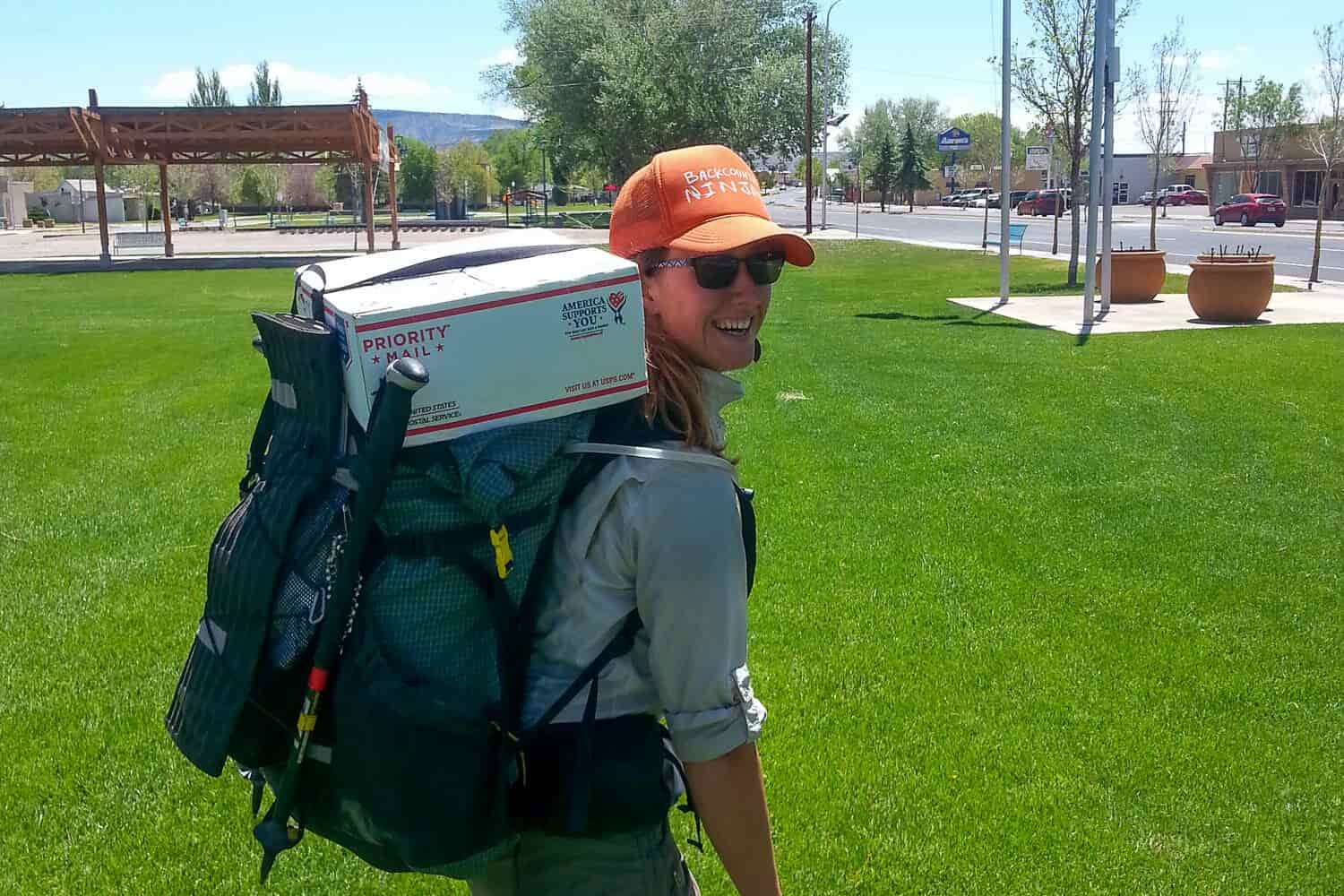 CDT thru-hiker in a town with a USPS box propped on top of her backpack. This box has resupply food for her to bring on the trail.