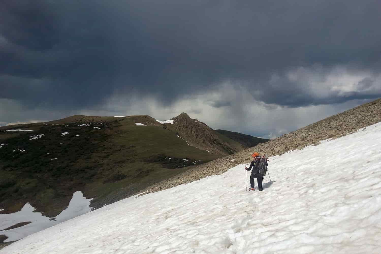 CDT backpacker walking across a snow field in the mountains with cloudy skies above.