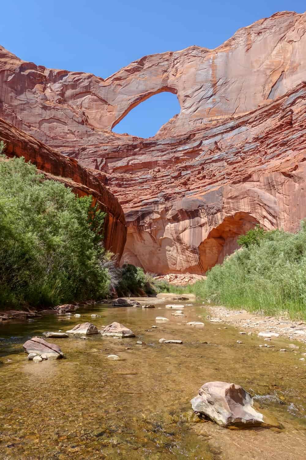 a stream flows below a large red rock wall with a hole in the wall above it where you can see blue sky