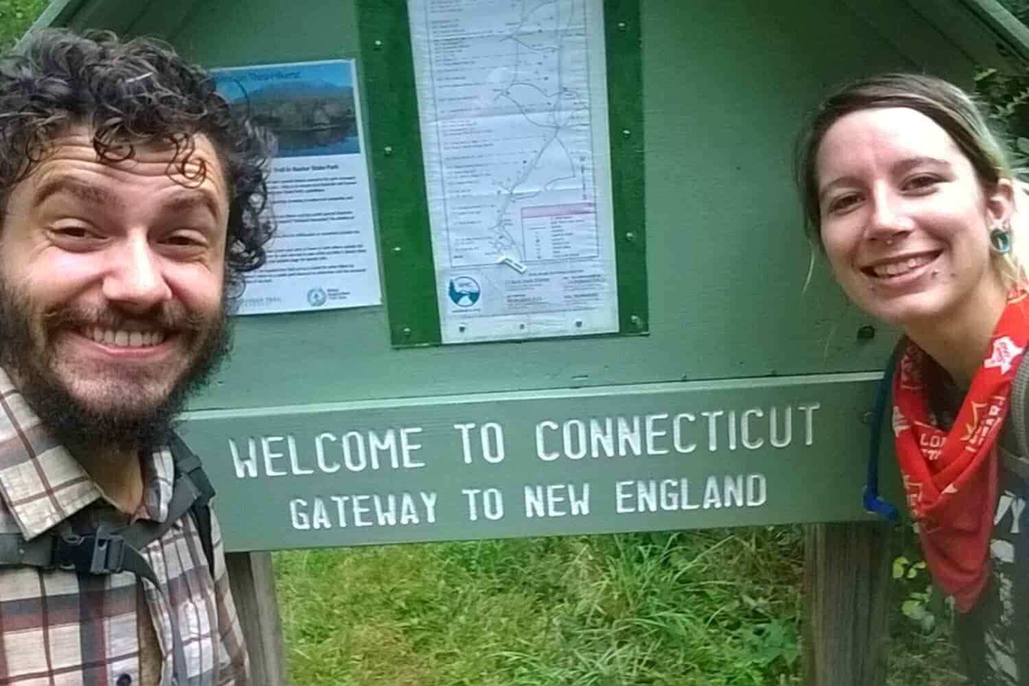 two Appalachian Trail thru-hikers take a photo in front of a sign that says "welcome to connecticut, gateway to new england"