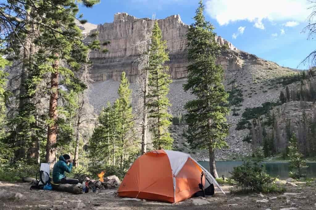 A male camper sits next to a campfire and orange tent near an alpine lake