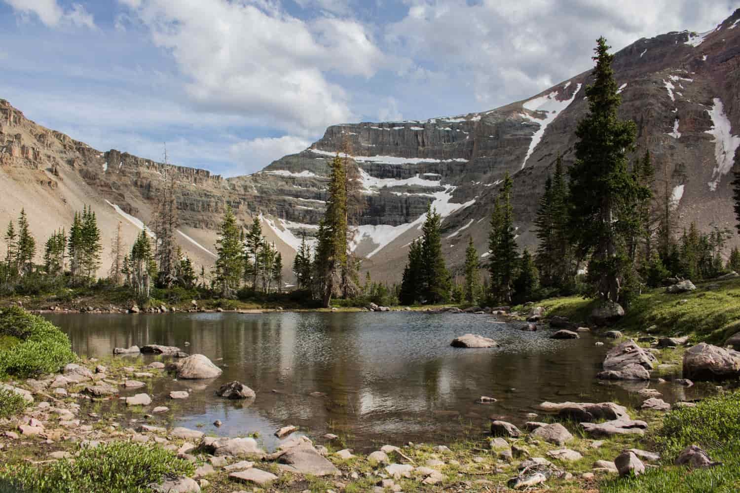 A small lake in a mountain basin surrounded by pines with patches of remnant snow along the peaks