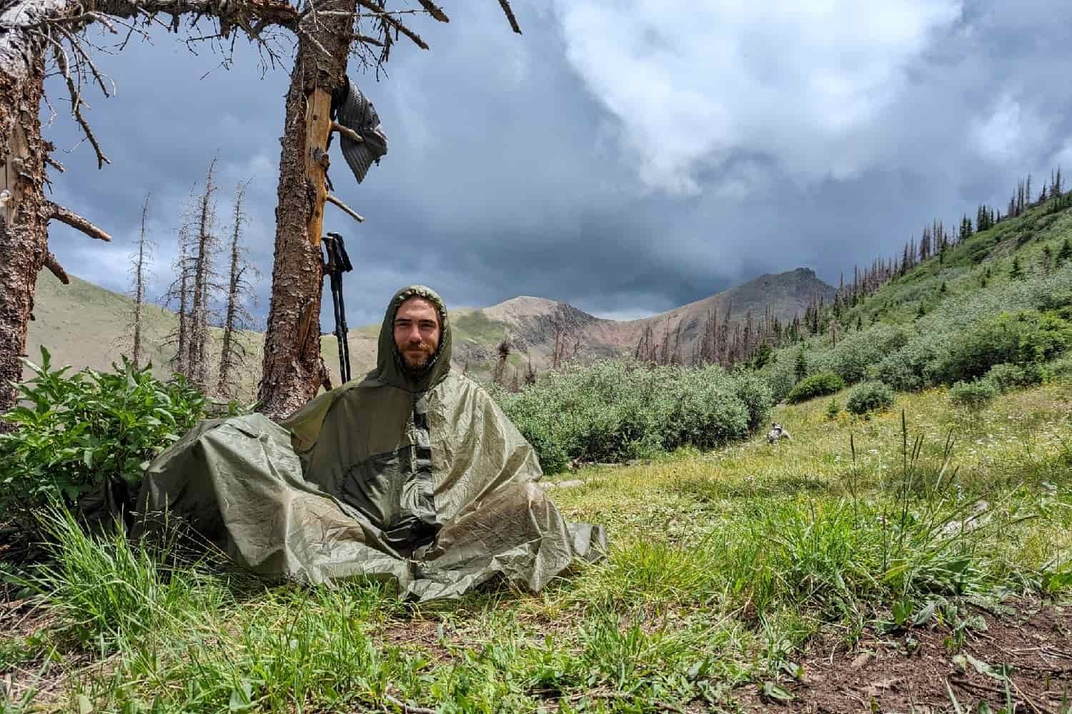 backpack seated on the ground in a rainstorm with a rain poncho on under a tree.