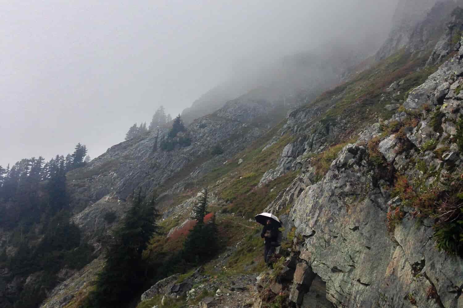 thru-hiker on the the PCT uses an umbrella on the rocky, cloud-covered trail.