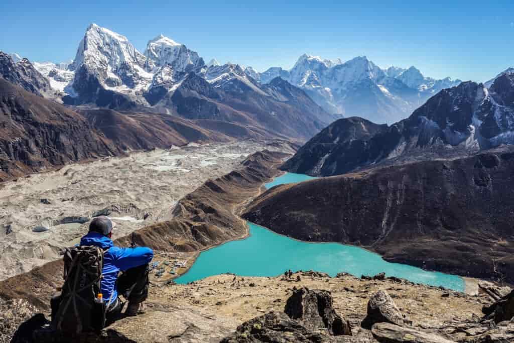 A hiker sits on a rock ledge looking down on a turquoise lake with angular mountains in the background