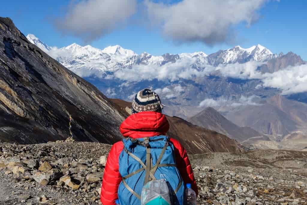 A hiker looks out on a rocky plateau with rugged, snowcapped mountains in the distance