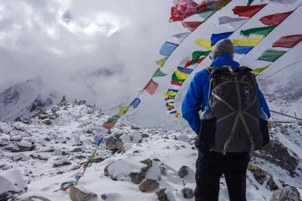 A backpacker stands underneath colorful flags while standing on top of a snowy mountain pass in Nepal