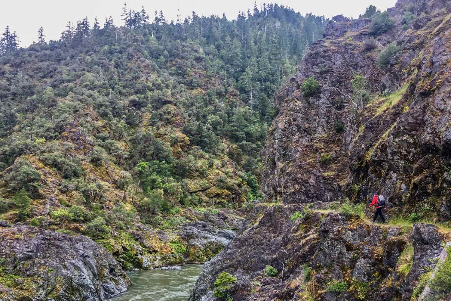 backpacker on a narrow ledge that wraps above a cliff with the Rogue River below on a cloudy, wet day.