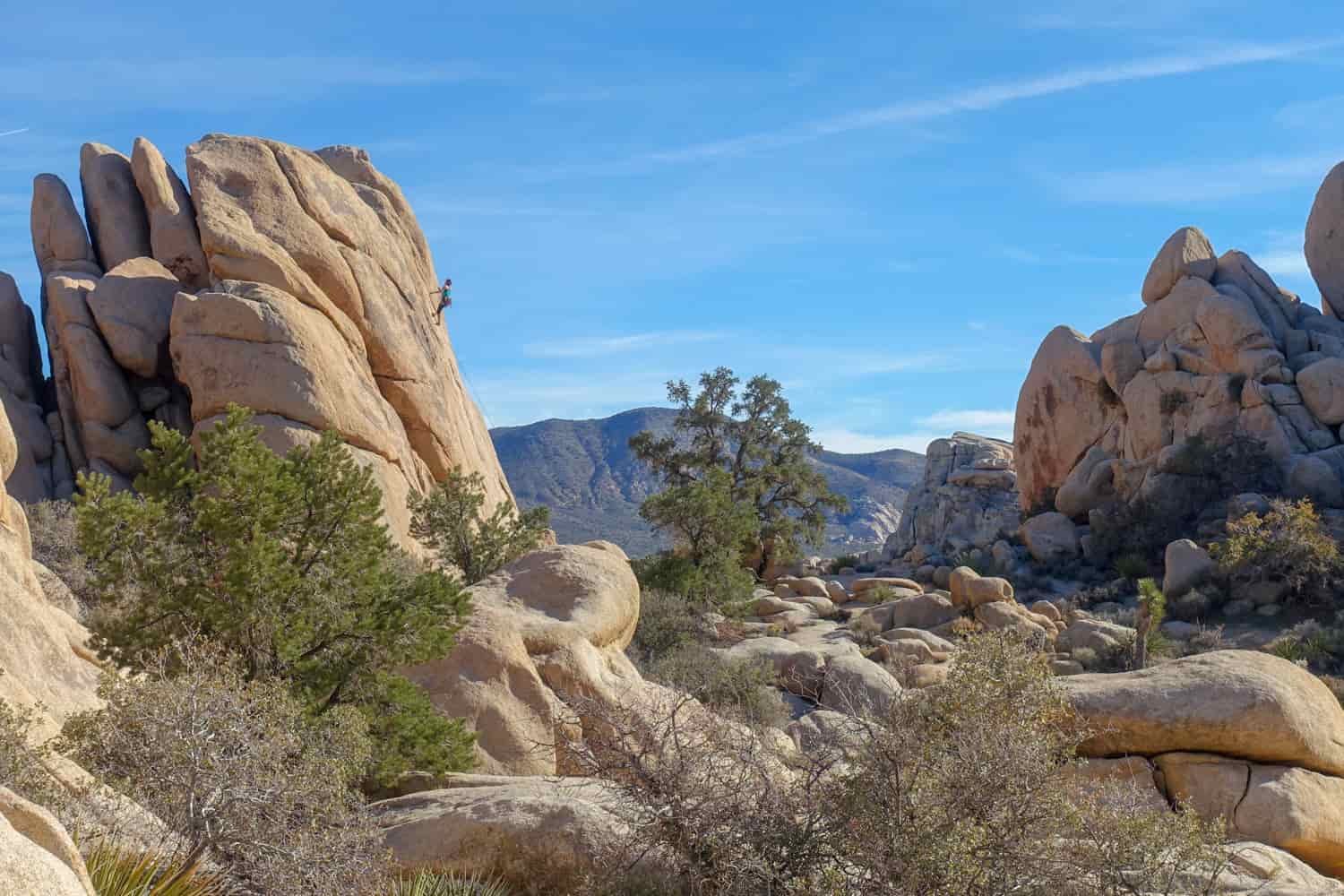 A valley full of massive boulders and rock promontories, including one with the small figure of a female climber