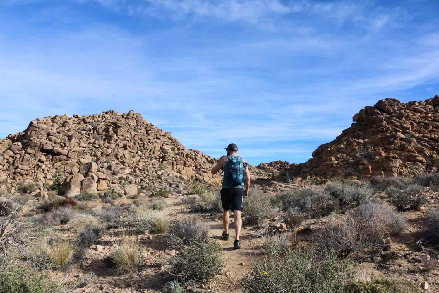 A male hiker in shorts walks through a desert arroyo toward a gap between hills covered in boulders