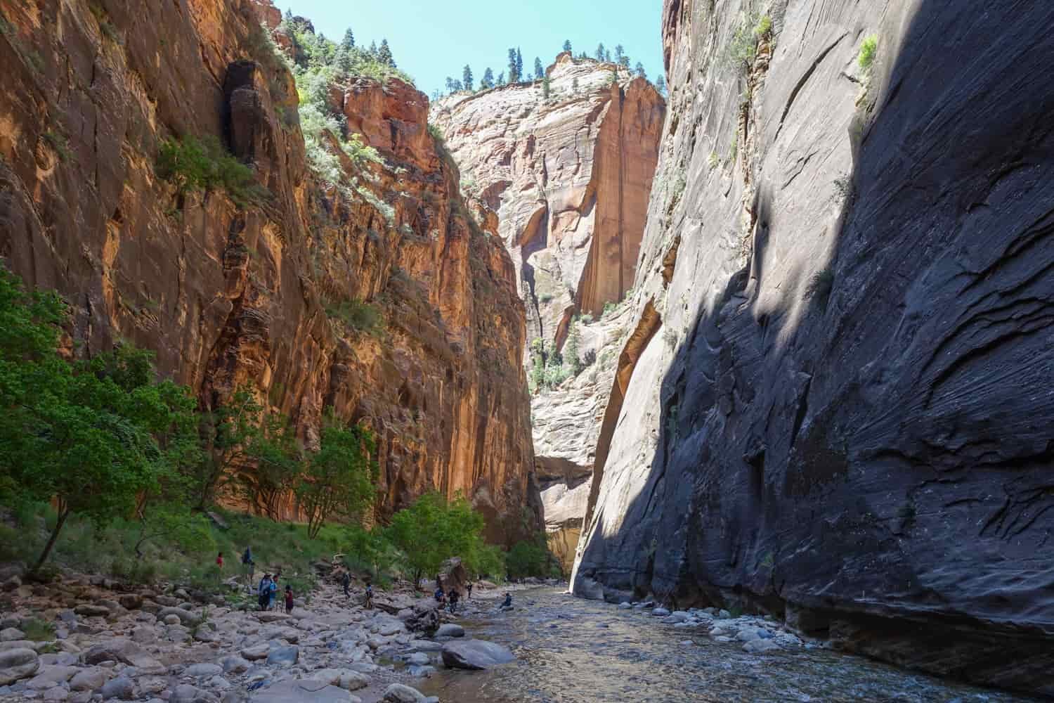 the narrows hike with people on the river bank covered in rip rap with orange sandstone walls rising on either side of the river