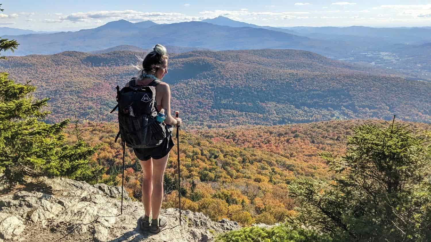 Backpacker on the long trail on a rock ledge overlooking rolling mountains in fall foliage.