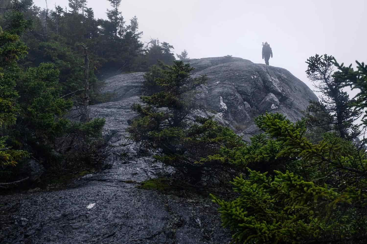backpacker approaching a mountain top on vermont's long trail. the trail is a rock slab with evergreen on either side.