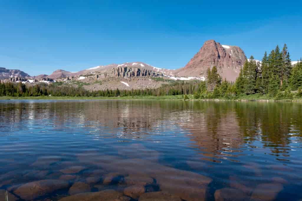 Underwater boulders visible beneath the surface of a mountain lake with pines and brown peaks in the background