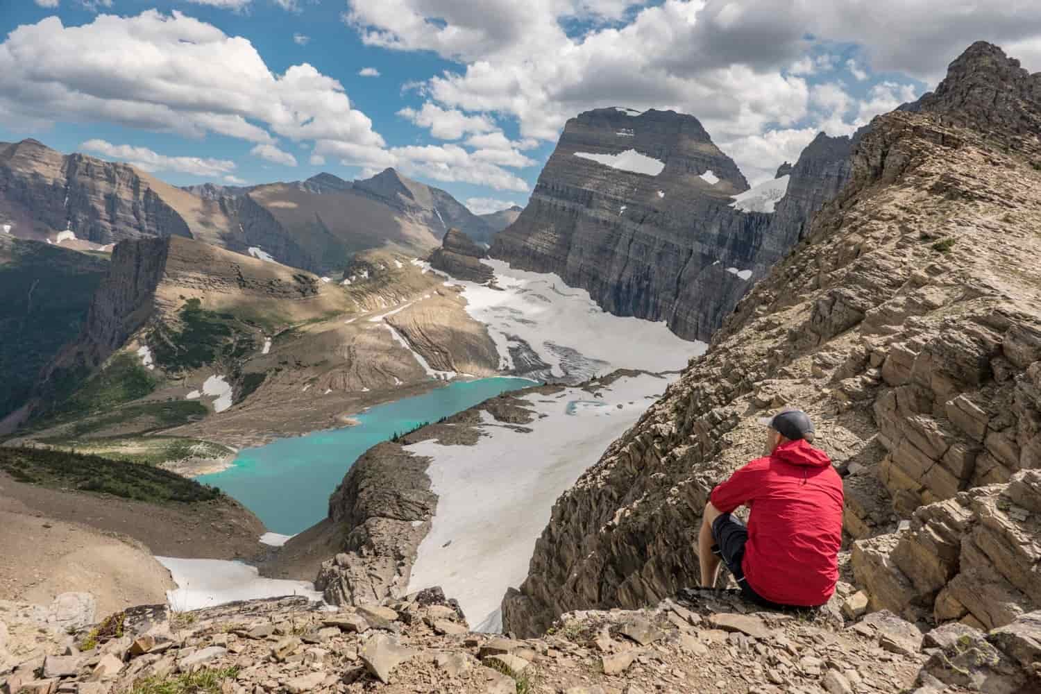 hiker seated on a rocky ledge overlooking a glacial tarn surrounded in mountains and snow in glacier national park