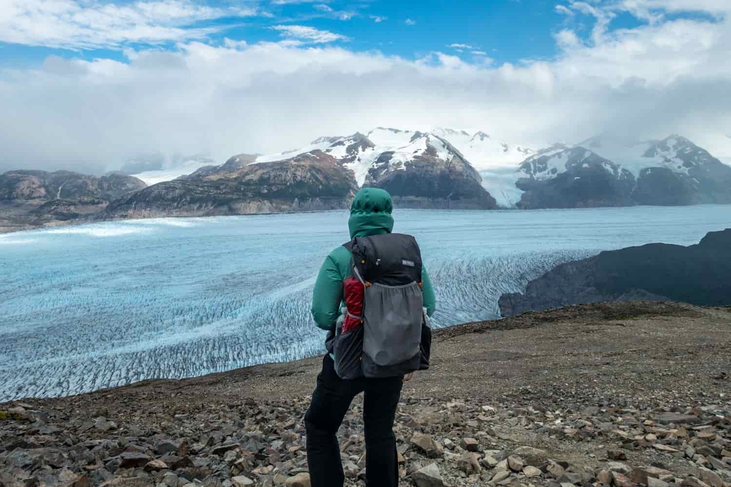 A hiker in a green jacket and a backpack looks out at a vast glacial landscape with snow covered mountains in the distance
