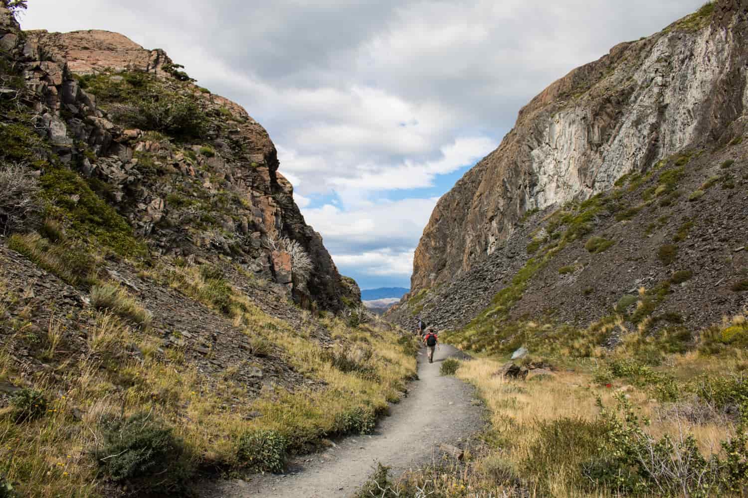 Two hikers walk down a well-graded path with rocky foothills on either side.