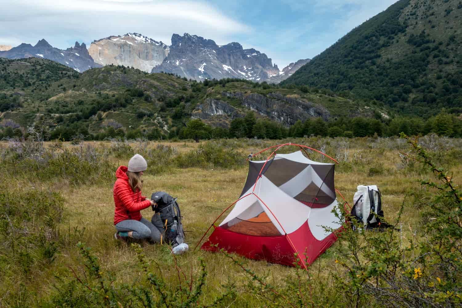A female backpacker looks in her backpack at a camp site surrounded by mountains
