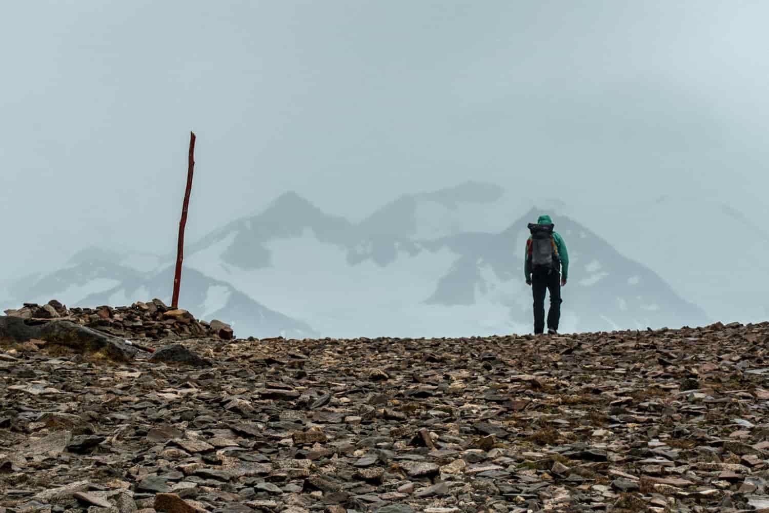 A hiker walks over a scree-covered saddle with snowy mountains ahead barely visible through a cloud