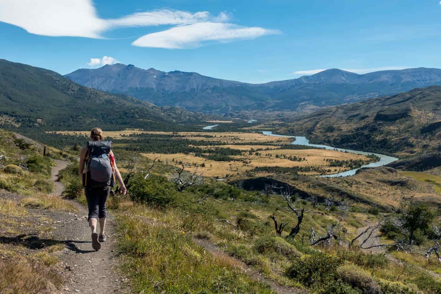 Rear view of a female hiker heading down a trail toward a river valley in the mountains