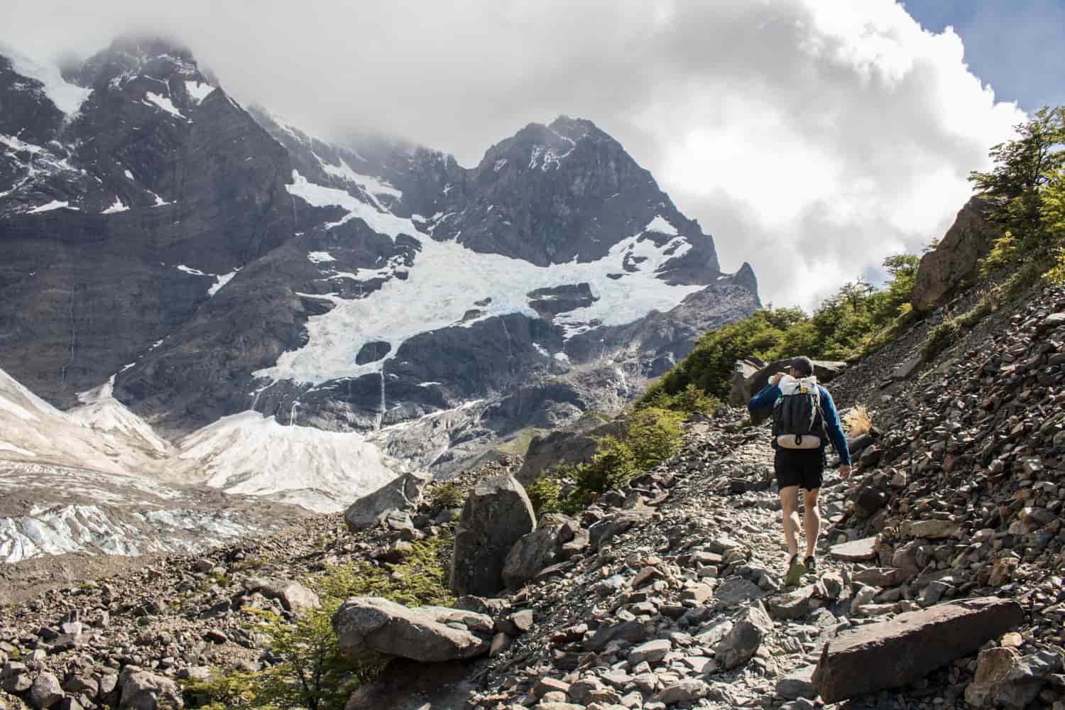 A backpacker sips from a water bottle while climbing a rocky trail that looks out over snowy mountains
