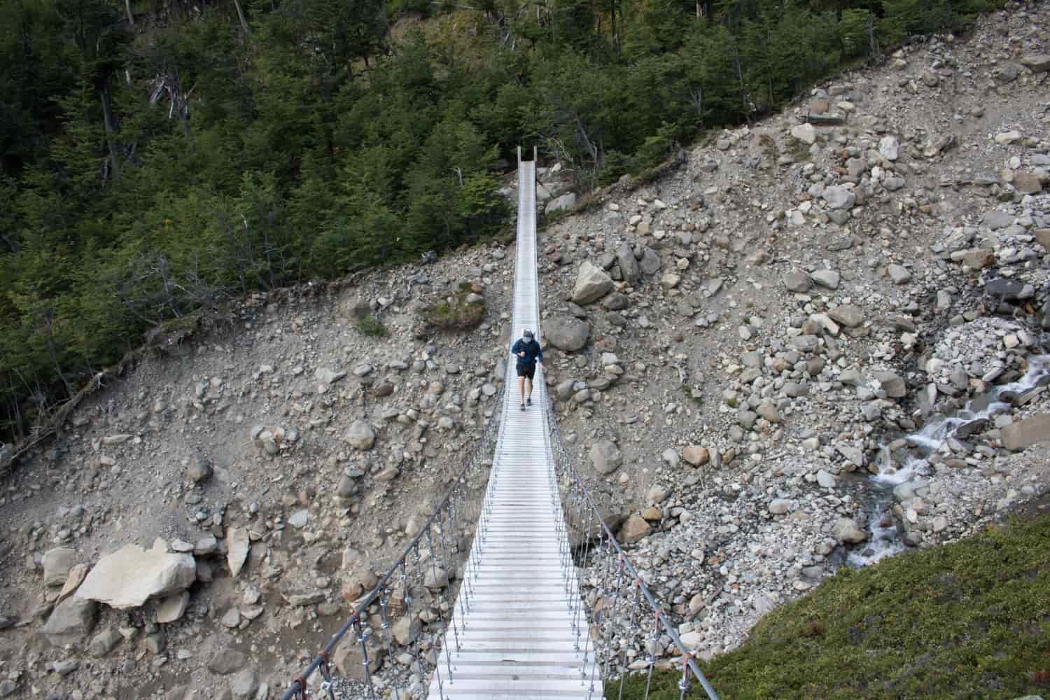 A hiker approaches on a suspension bridge over a ravine with snowmelt flowing down