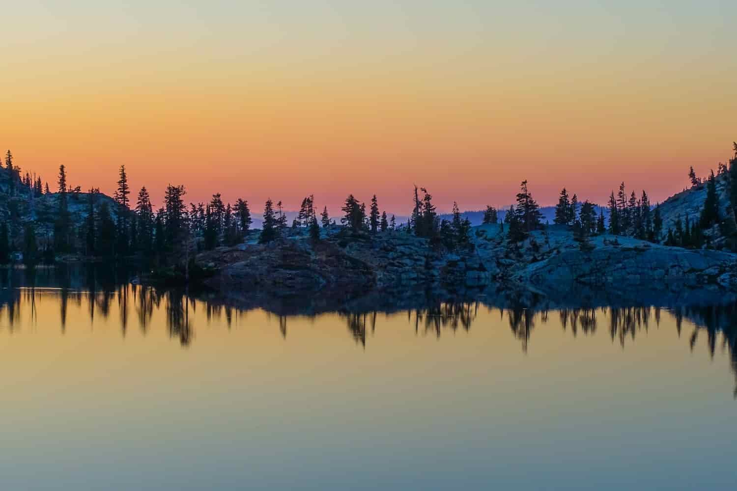 orange, yellow and pink sunset on an alpine lake in the trinity alps wilderness with the trees and sunset reflected on the lake