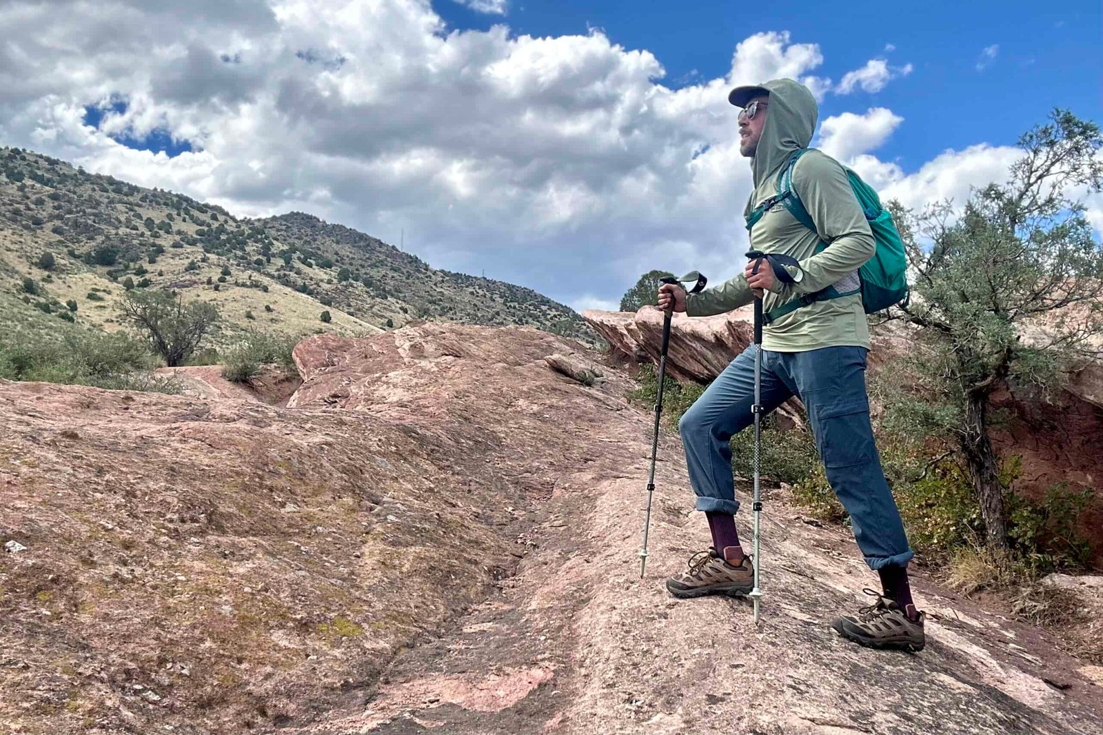 Man wearing a green sun hoody, backpack, pants, and Merrell Moab 3 poses on an uphill hike as he takes in the view of rocks and shrubs.