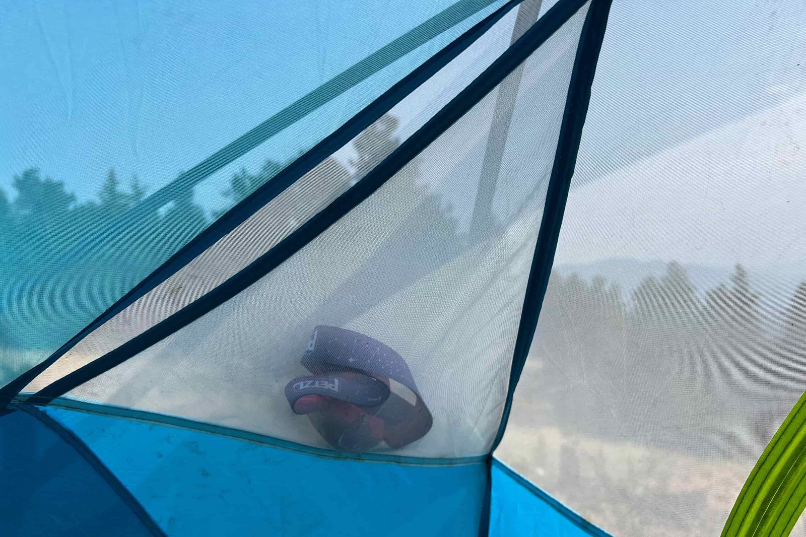 The image shows an interior mesh pocket on the Nemo Aurora Highrise 4 contaiing a headlamp. Through the tents mesh you can see pine trees in the distance.