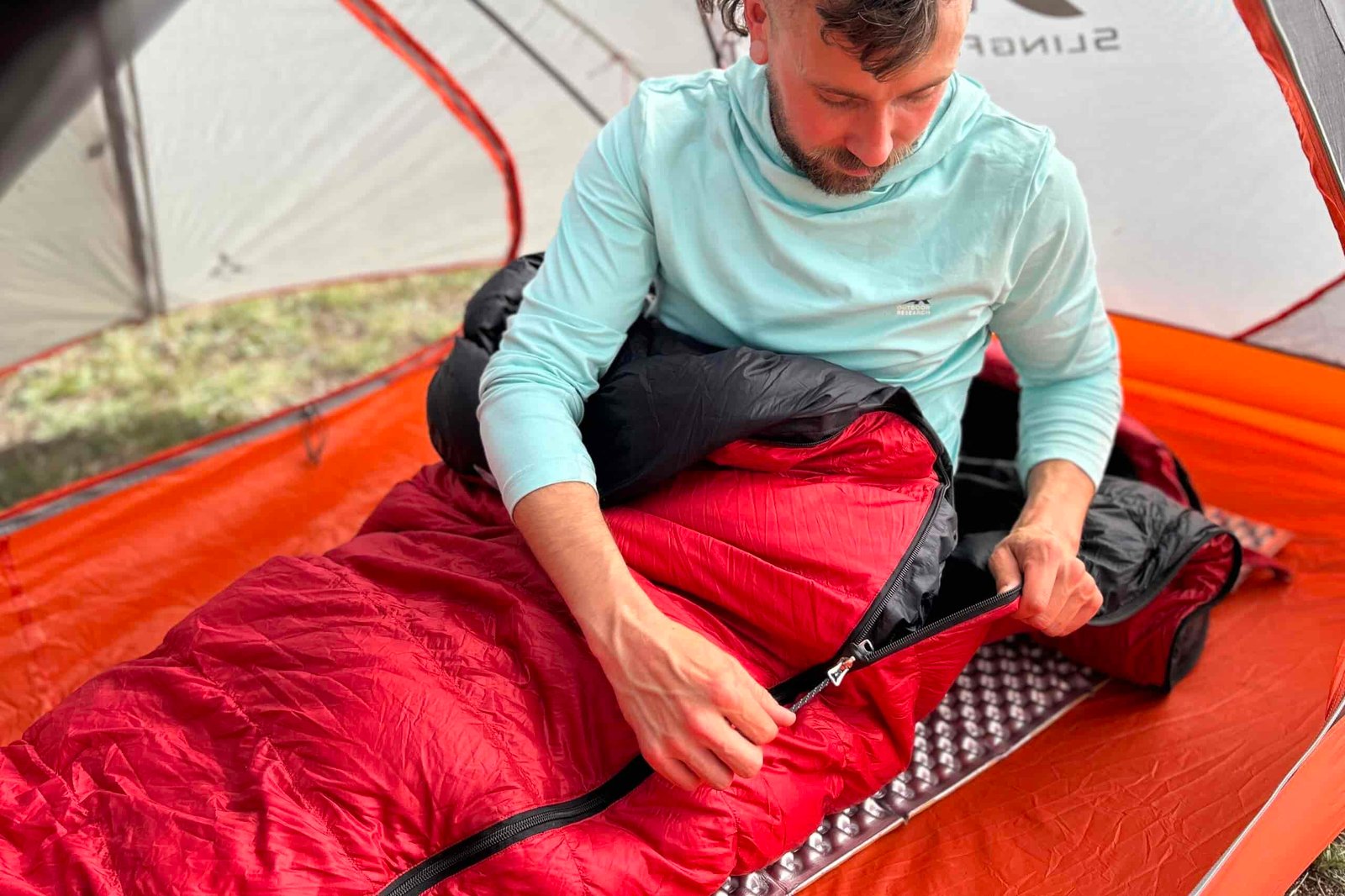 A man sits up on a pad and is in a sleeping bag from the waist down. He messes with the zipper on a red sleeping bag inside of a tent.