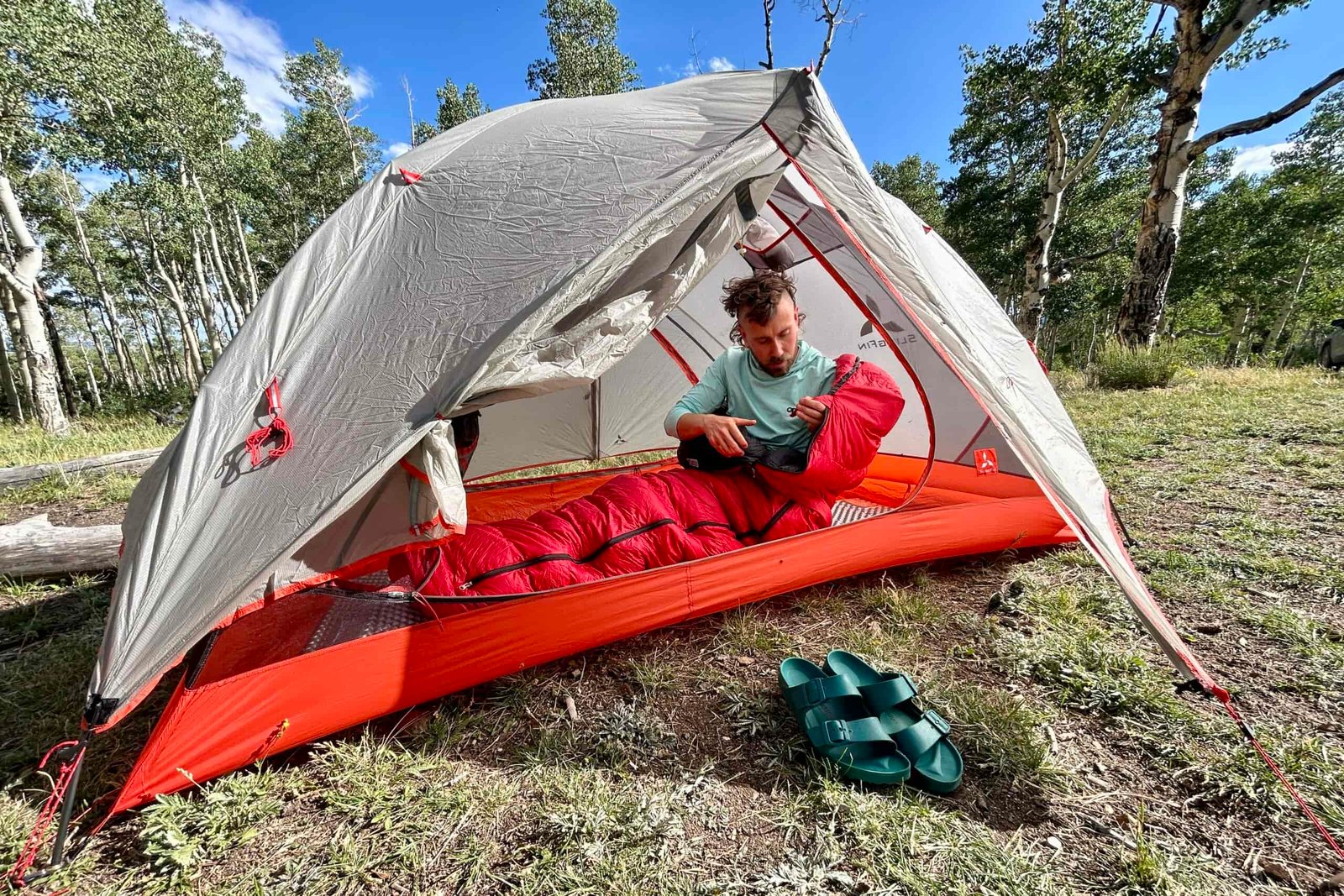 A man messing with the zipper on a red sleeping bag inside of a tent on a beautiful bluebird day. Image is from outside t he tent.