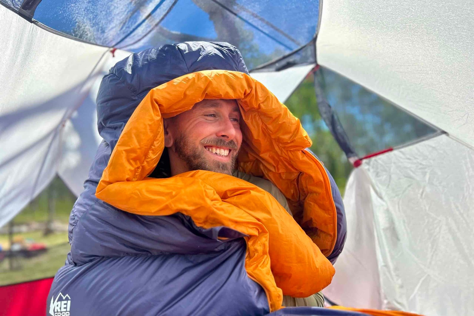 A man sits up in a sleeping bag with the hood over his head. He is smiling and looking off camera.