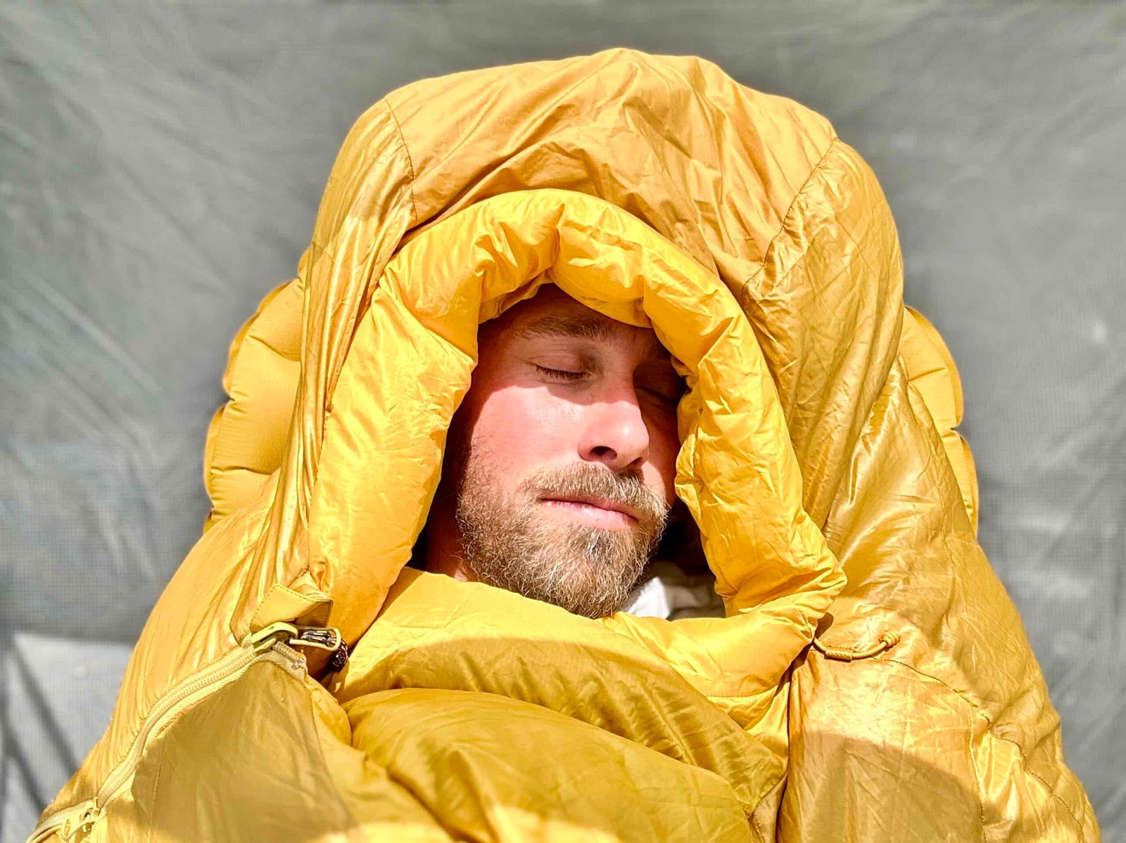 Aclose up image of a man sleeping in a sleeping bag, with morning sun on his face. The hood is cinched around his face and it looks very cushy and plush.