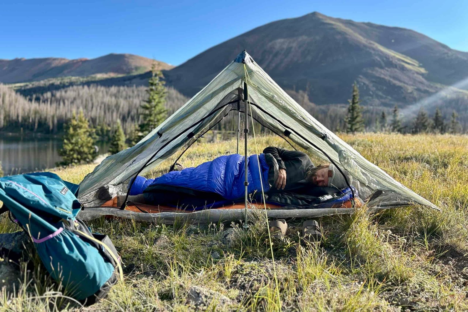A man sleeping in a tent in a blue sleeping bag with his backpack on the ground next to his tent.