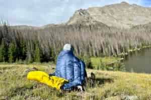 A man sits with a sleeping quilt wrapped around his back looking over an alpine lake with mountains behind it.