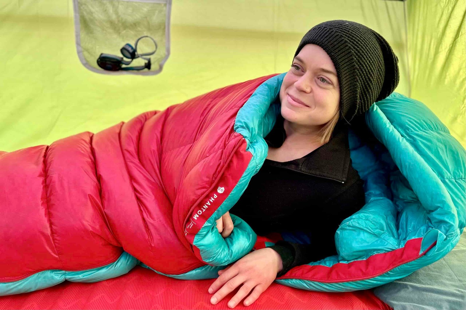 A woman in a sleeping bag sits up on one arm and looks into the distance.