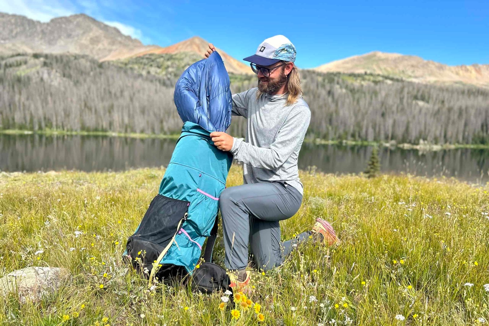 A man pulls a down quilt out of a backpack while kneeling in a field of flowers with a lake and mountains behind.