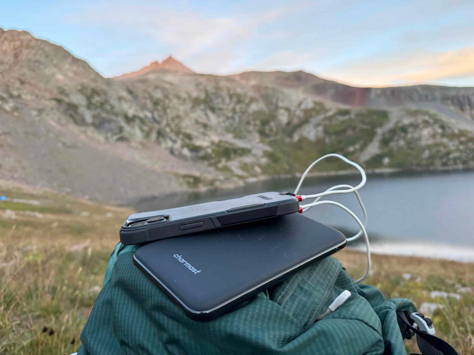 Close up of the Charmast power bank charging a phone that is sitting on a backpack with a lake and mountain in the background.