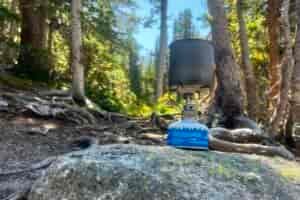 A low angle view of the Snow Peak Gigapower 2.0 sitting on a rock with trees in the background. The stove is installed with a small fuel canister, and a small cook pot is on the pot stand.
