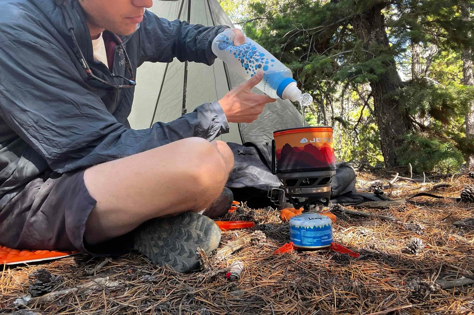 A hiker filters water into his MiniMo pot. The pot is installed with its burner, fuel canister, and canister support. The background has trees and backpacking gear.