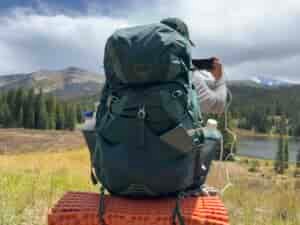 A hiker has their back to the camera while taking a photo. The phone is hooked up to the UGREEN charger in the back pocket of the pack. There are trees and a mountain lake in the background.