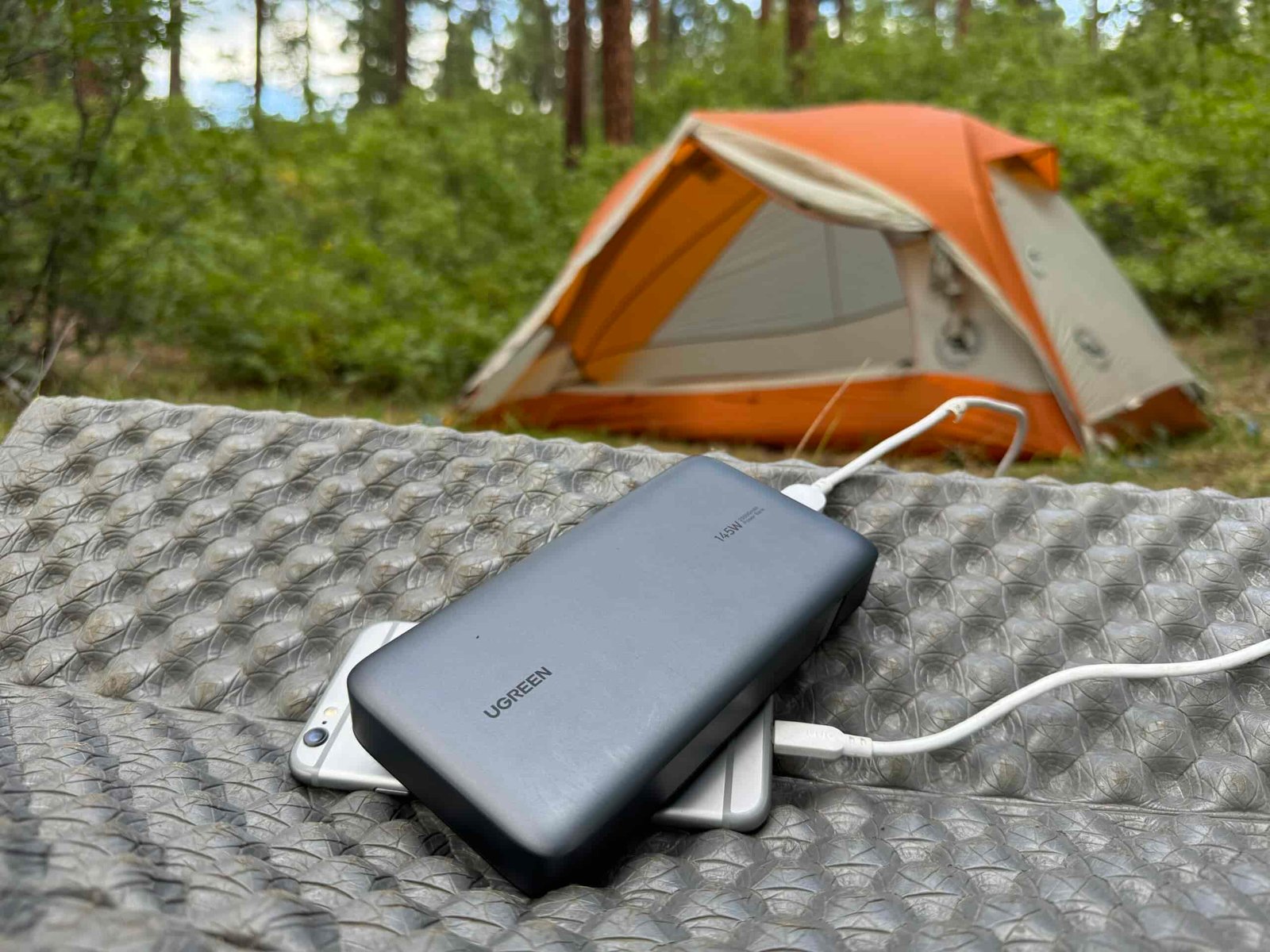 The UGREEN and a phone sit in front of a tent in the woods.