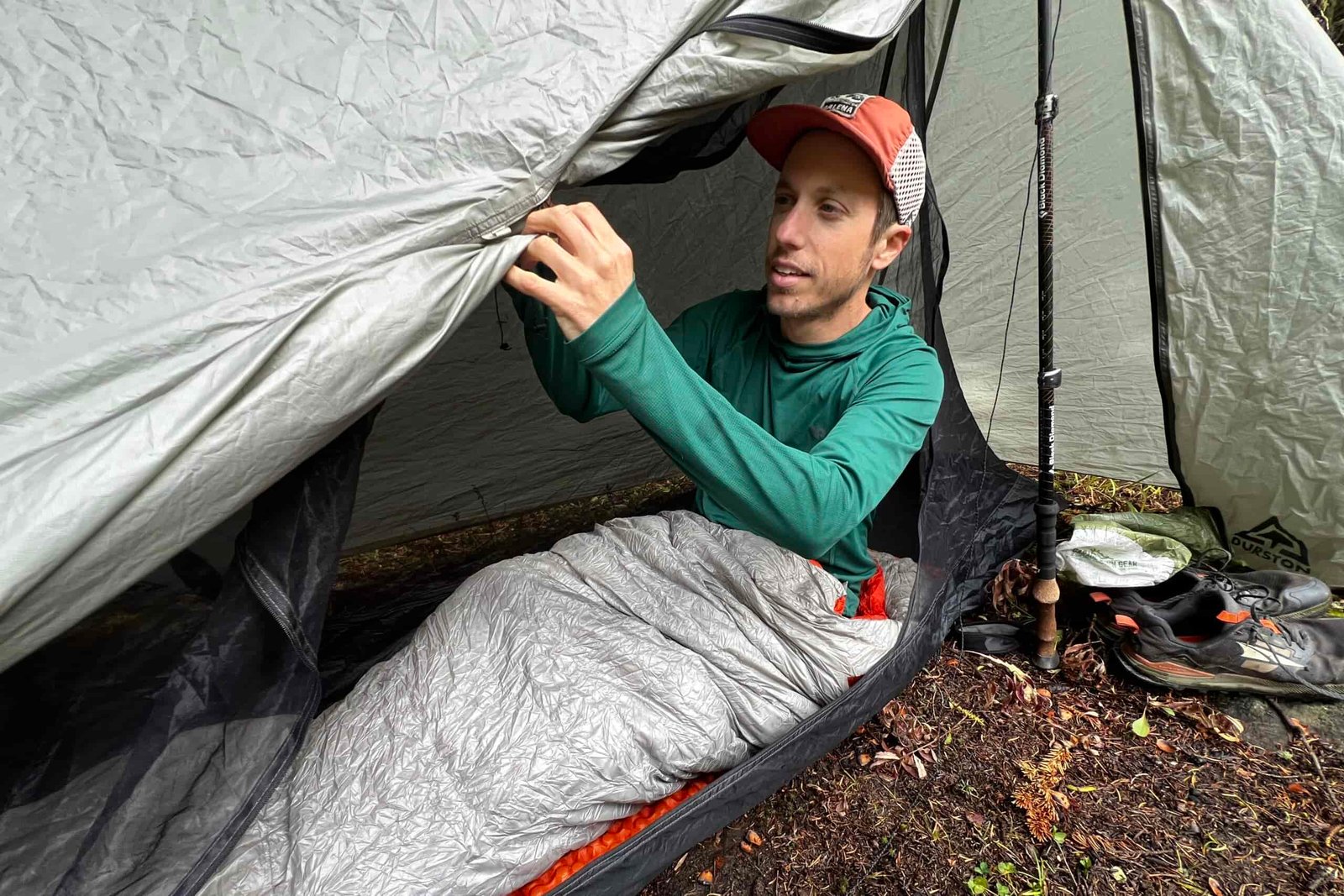 A hiker adjusts his tent while sitting in a sleeping bag, wearing the Outdoor Research Echo Hoodie.