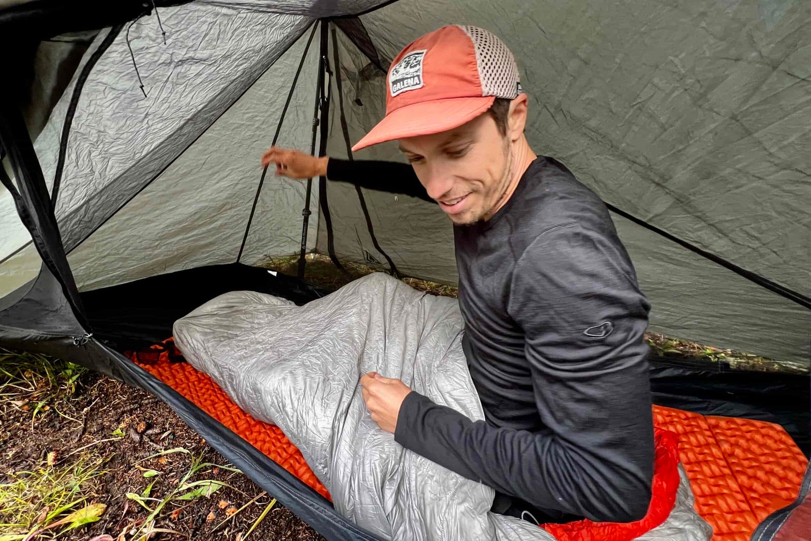 A hiker sits in his sleeping bag in a tent while wearing the Icebreaker Merino Oasis 200 base layer. Tent walls are in the background.