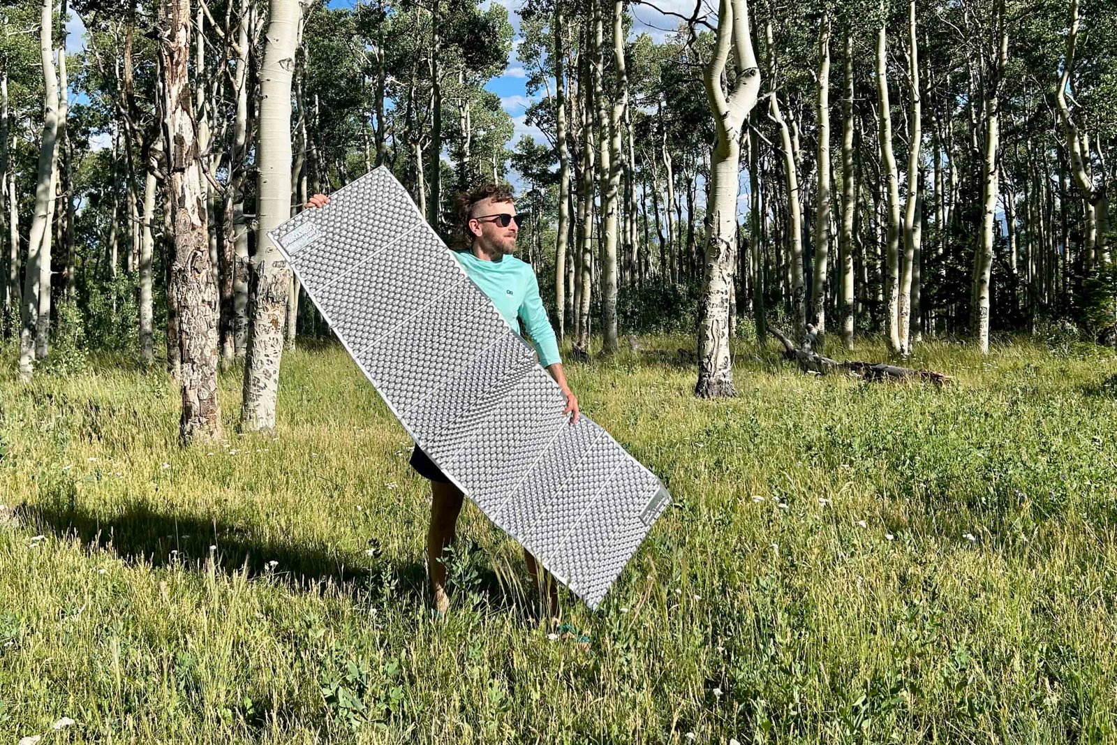 A man holds a grey mat in a grassy area in the woods.
