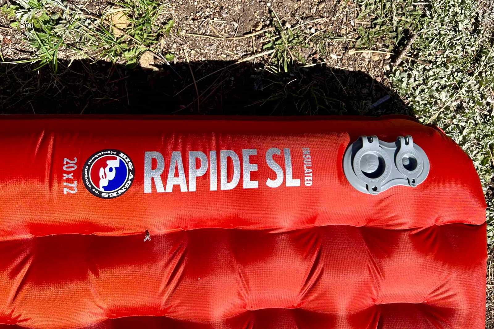 A red inflatable sleeping pad labeled "RAPIDE SL" lays on grass, featuring a Big Agnes logo, and dual air valves, with its quilted bottom visible.