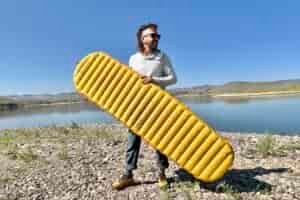 A man stands next to a lake gripping a large yellow inflatable camping pad.