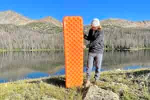 A man stands in front of a lake and mountains holding an orange inflatable mattress.